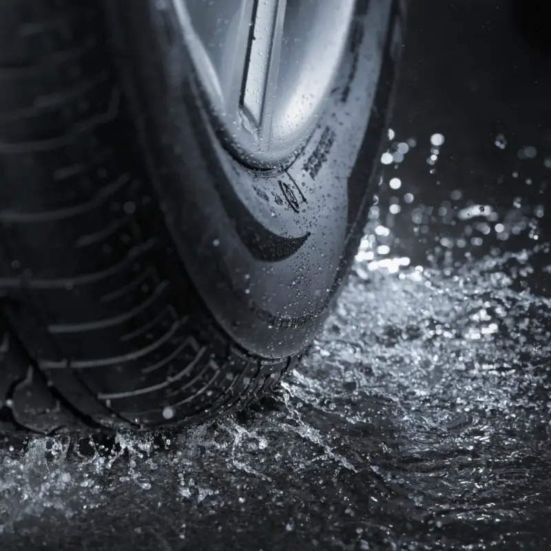 Close-up of a car tire and alloy wheel splashing through water on wet pavement.