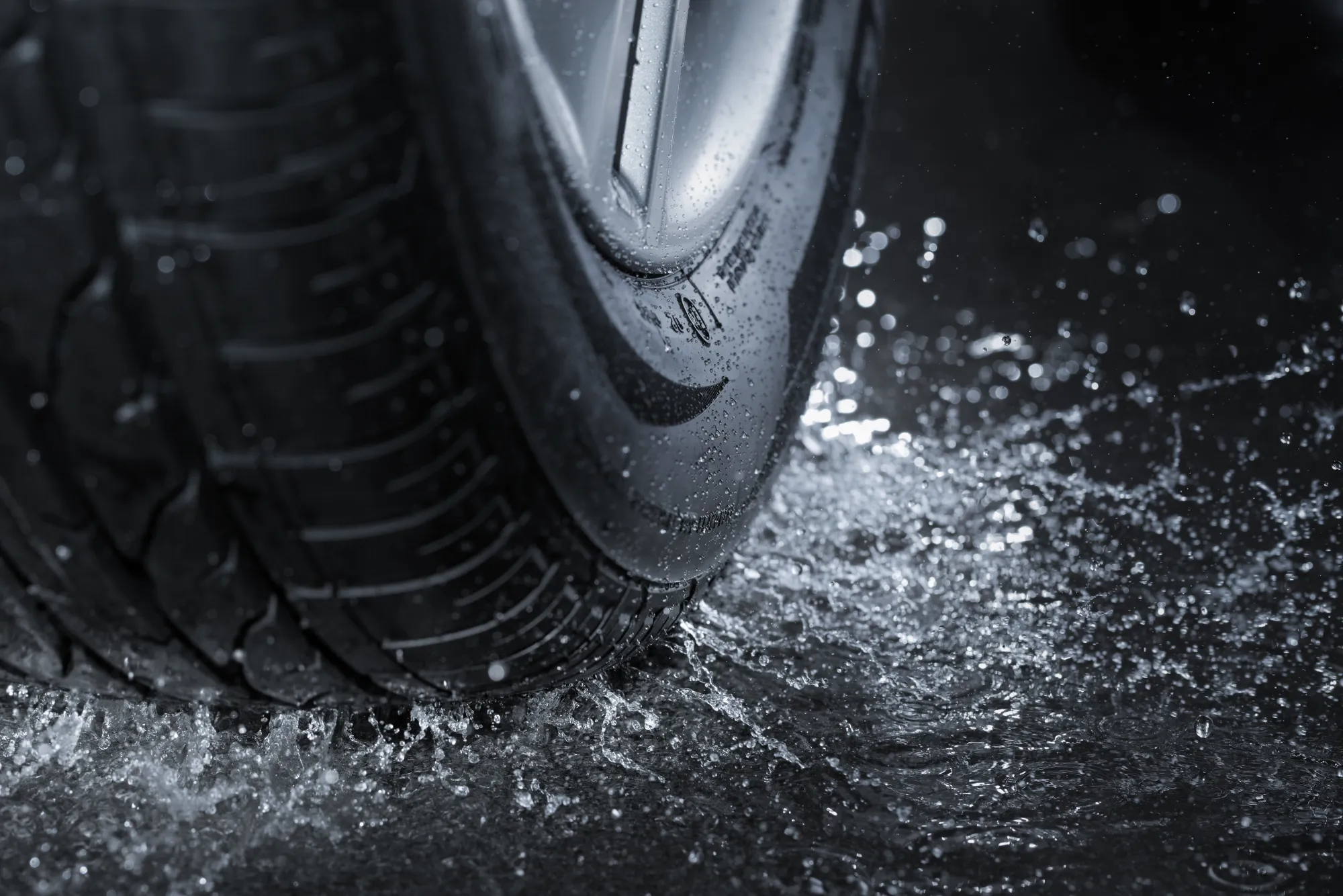 Close-up of a car tire and alloy wheel splashing through water on wet pavement.