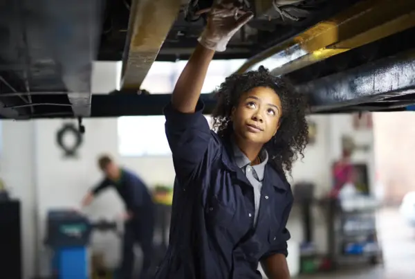 A female mechanic in navy coveralls reaches up to inspect the underside of a car raised on a lift inside an auto repair shop.