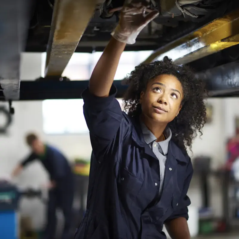 A female mechanic in navy coveralls reaches up to inspect the underside of a car raised on a lift inside an auto repair shop.