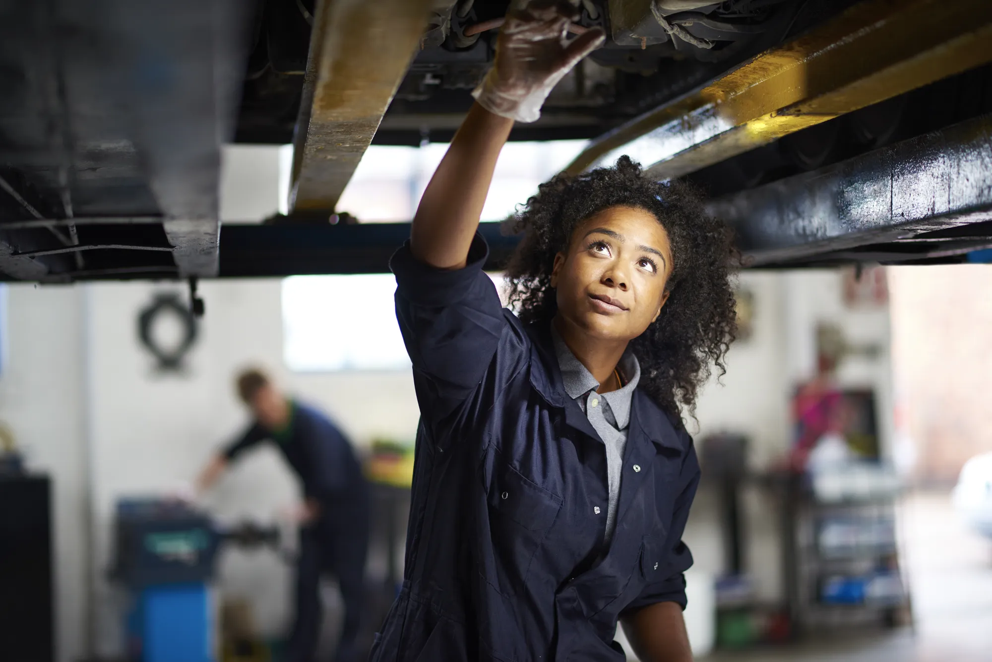 A female mechanic in navy coveralls reaches up to inspect the underside of a car raised on a lift inside an auto repair shop.