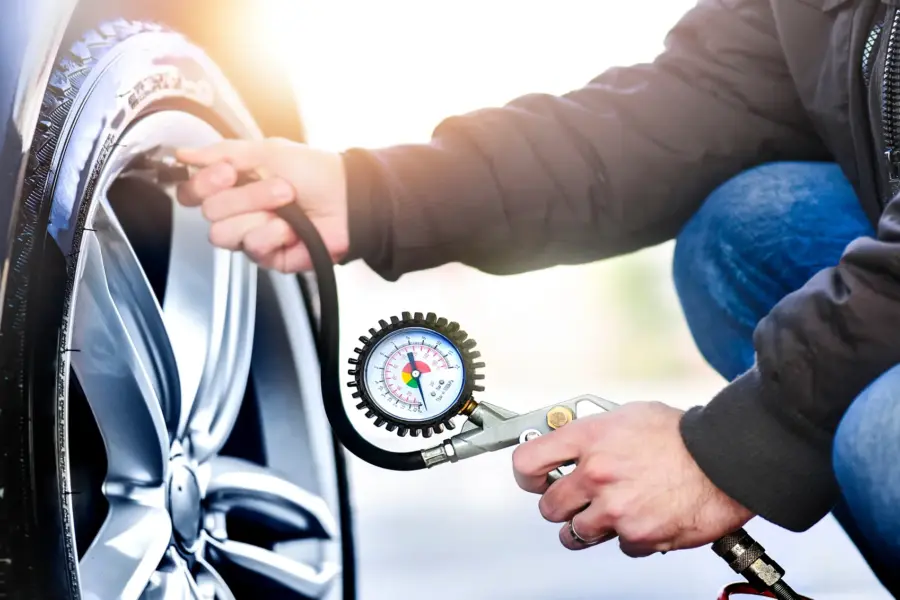 A man kneeling next to a car wheel checks the tire's air pressure with a hose-connected pressure gauge.