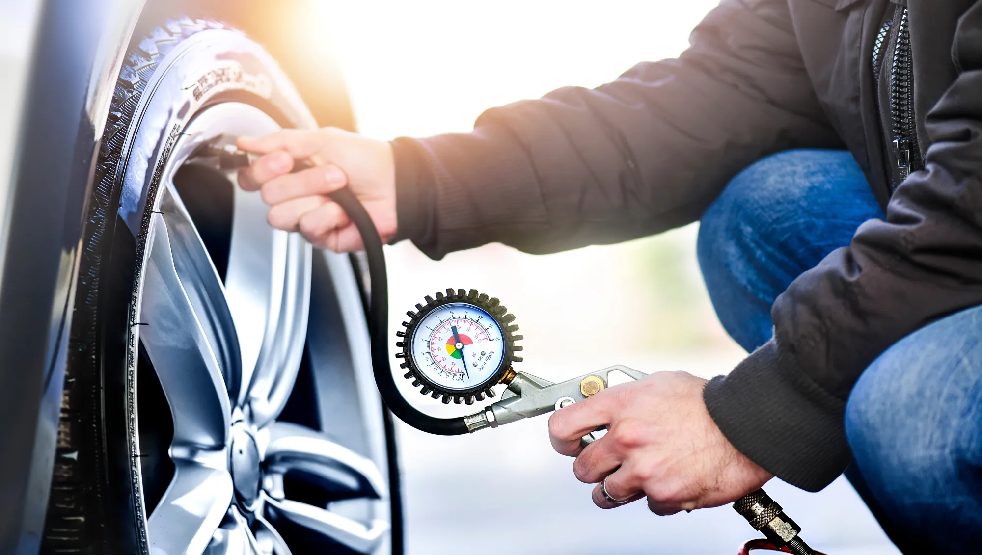 A man kneeling next to a car wheel checks the tire's air pressure with a hose-connected pressure gauge.