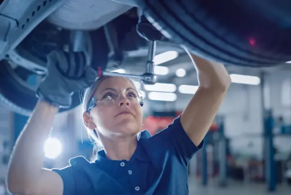 A female mechanic in safety glasses reaches up with a ratchet to work under a car raised on a lift in an auto repair shop.