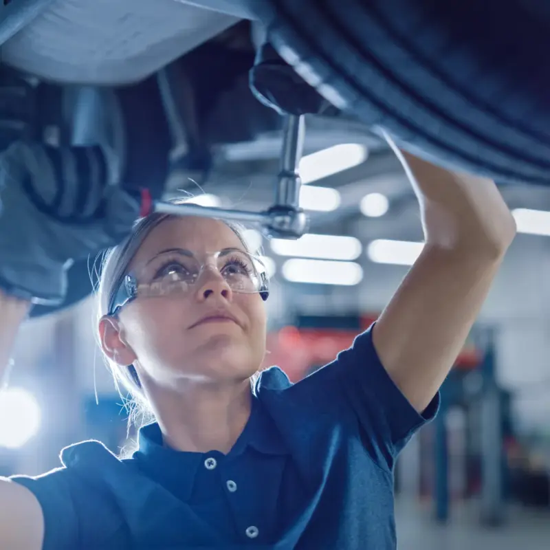 A female mechanic in safety glasses reaches up with a ratchet to work under a car raised on a lift in an auto repair shop.
