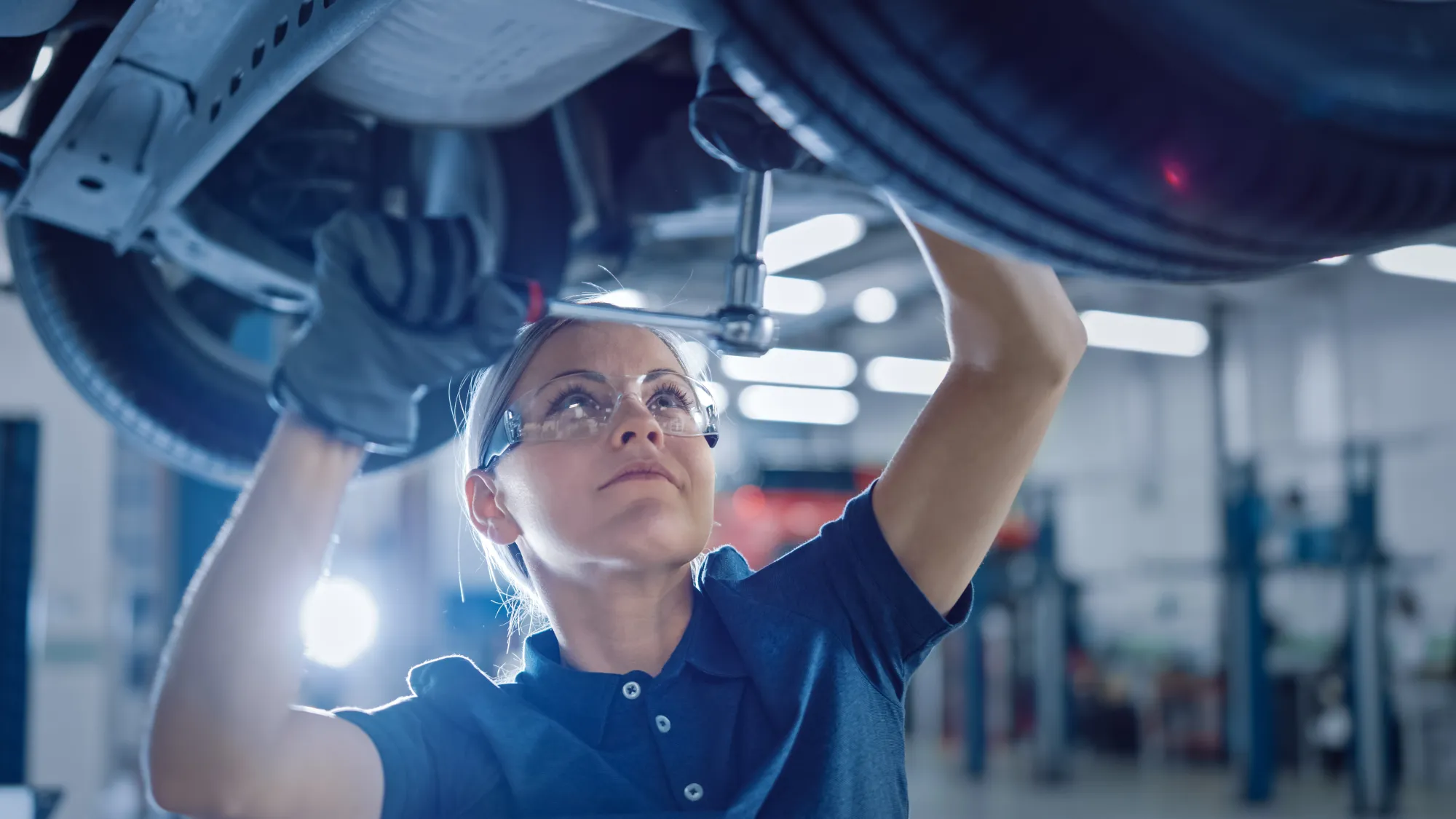 A female mechanic in safety glasses reaches up with a ratchet to work under a car raised on a lift in an auto repair shop.