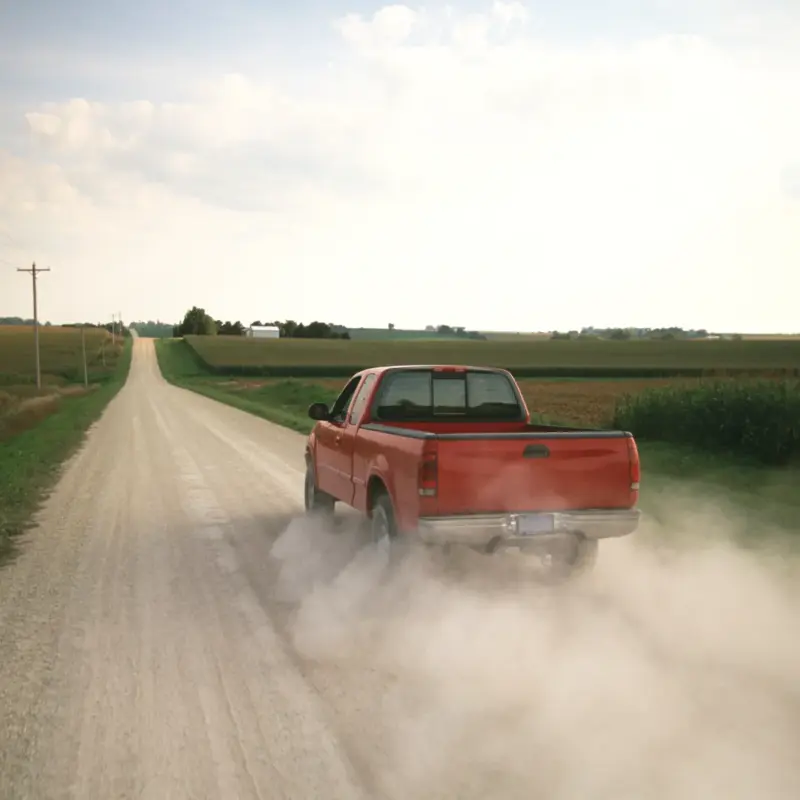 A red pickup truck driving away down a gravel road between cornfields, raising a large cloud of dust behind it.